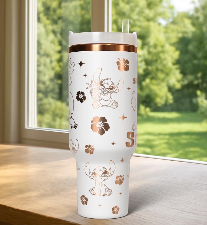 May include: White tumbler with a rose gold band and lid, featuring a repeating pattern of the character Stitch, flowers, and stars. The tumbler has a straw and the letter "S" on the side. The tumbler is sitting on a wooden surface.