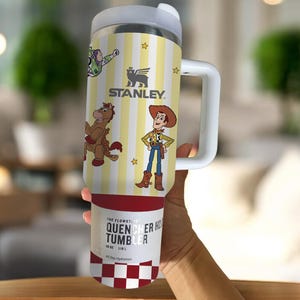 May include: A stainless steel Stanley Quencher H2.0 Flowstate tumbler with a white handle and lid. The tumbler features a yellow and white striped design with cartoon characters and the Stanley logo. The bottom of the tumbler has a red and white checkered pattern.