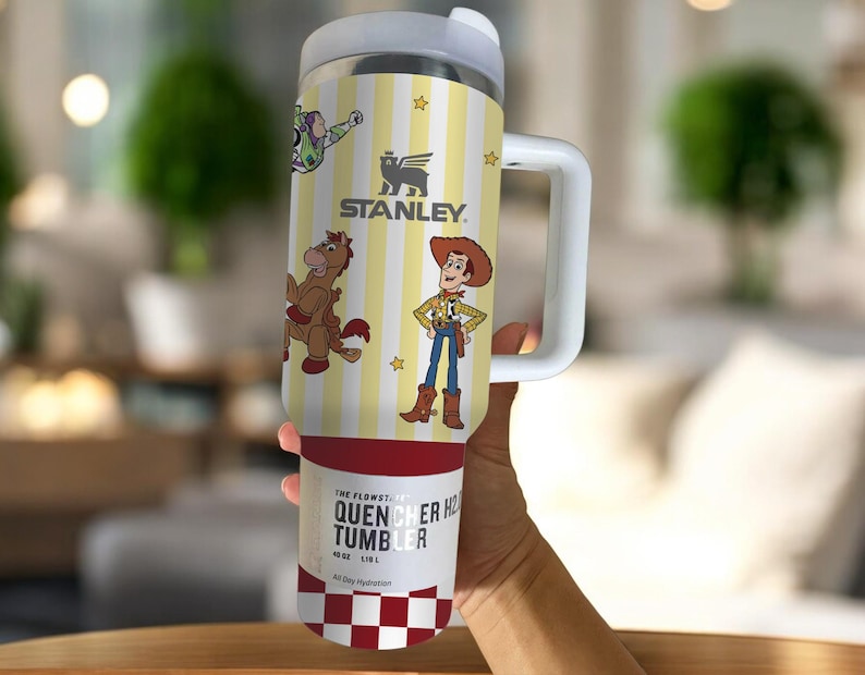 May include: A stainless steel Stanley Quencher H2.0 Flowstate tumbler with a white handle and lid. The tumbler features a yellow and white striped design with cartoon characters and the Stanley logo. The bottom of the tumbler has a red and white checkered pattern.