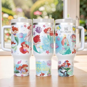 Puede incluir: Tres vasos Stanley blancos con ilustraciones de Ariel de La Sirenita de Disney. Cada vaso tiene un dise&ntilde;o diferente, incluyendo a Ariel, conchas marinas y criaturas marinas. Los vasos tienen asas y tapas blancas.