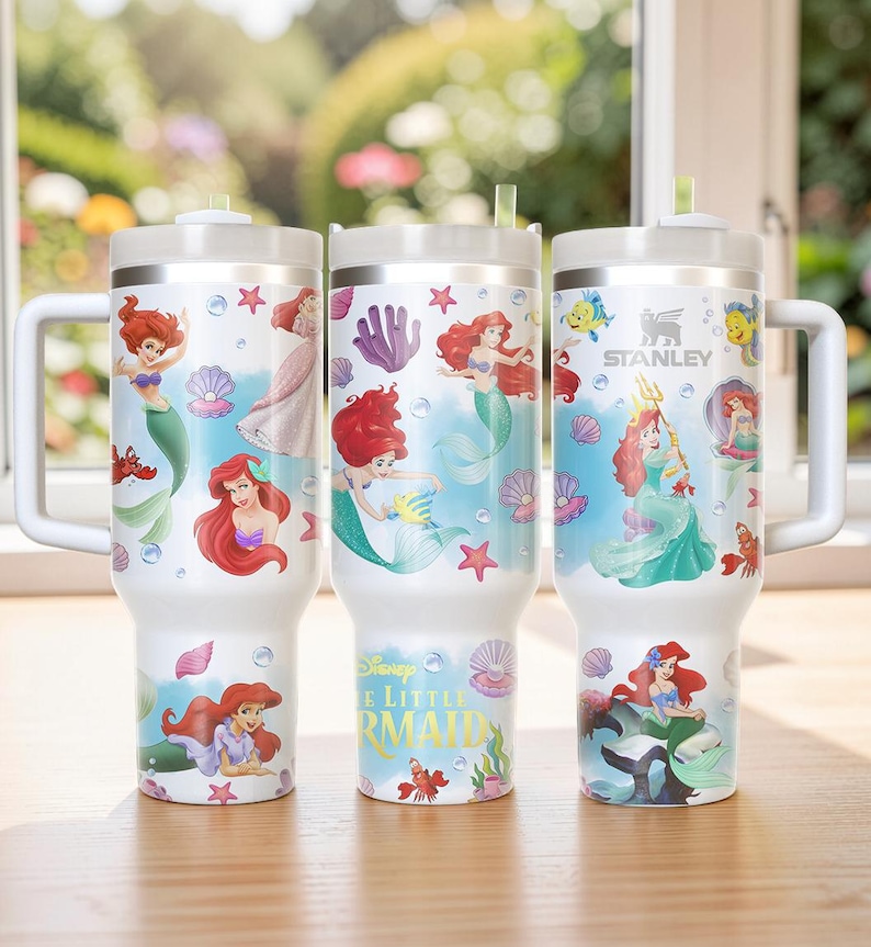 Puede incluir: Tres vasos Stanley blancos con ilustraciones de Ariel de La Sirenita de Disney. Cada vaso tiene un dise&ntilde;o diferente, incluyendo a Ariel, conchas marinas y criaturas marinas. Los vasos tienen asas y tapas blancas.