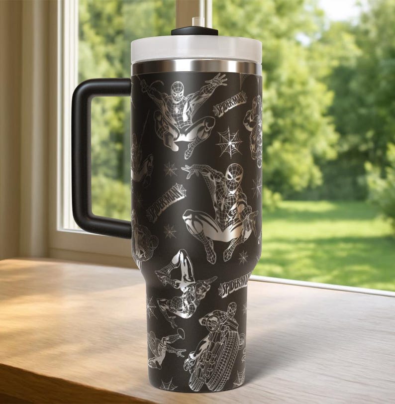 Pu&ograve; includere: Tazza da viaggio nera con coperchio e manico bianchi, con un motivo ripetuto di illustrazioni di Spider-Man e la parola "SPIDERMAN" in argento. La tazza ha un manico grande e un bordo argentato.