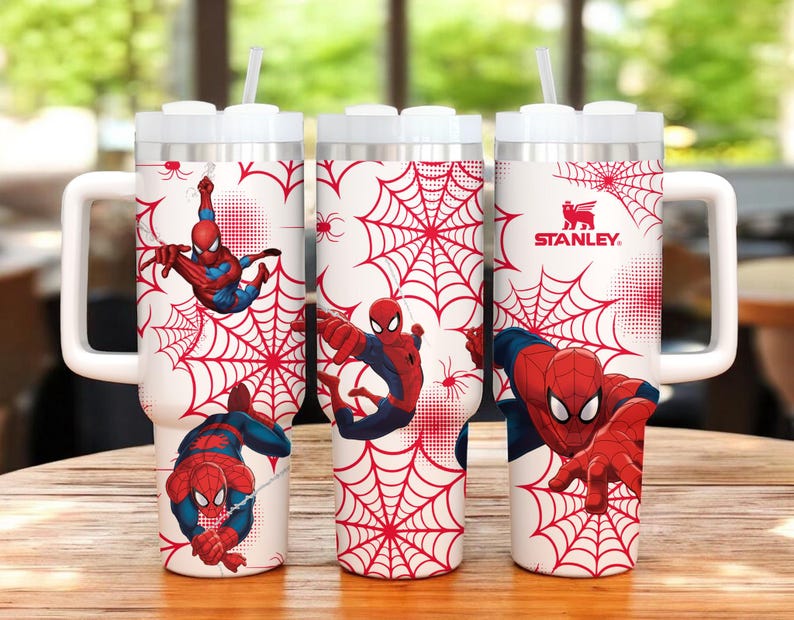 May include: White Stanley tumbler featuring Spider-Man graphics. The design includes red spiderwebs and illustrations of Spider-Man in action. The tumbler has a white handle and lid, and a straw.