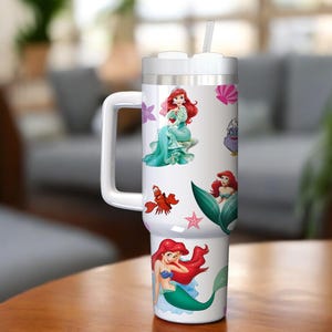 Puede incluir: Un vaso t&eacute;rmico blanco con asa y pajita, decorado con ilustraciones coloridas de una sirena y criaturas marinas. El vaso tiene una tapa plateada y un asa blanca.