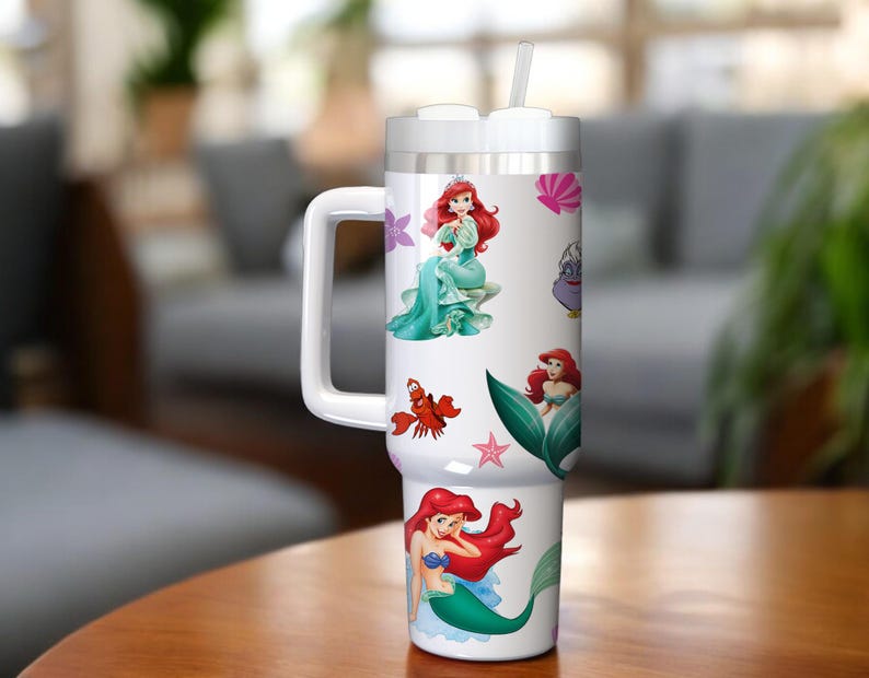 Puede incluir: Un vaso t&eacute;rmico blanco con asa y pajita, decorado con ilustraciones coloridas de una sirena y criaturas marinas. El vaso tiene una tapa plateada y un asa blanca.