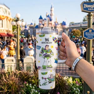 Pu&ograve; includere: Bicchiere Stanley bianco con Buzz Lightyear di Toy Story. Il bicchiere ha un manico e una cannuccia, con un design di Buzz Lightyear e altri elementi di Toy Story. Il logo Stanley &egrave; visibile.