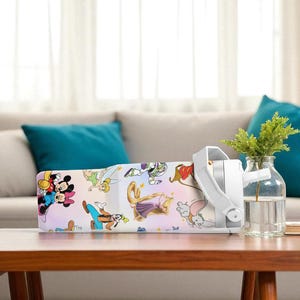 May include: A white air purifier featuring Disney characters, placed on a wooden table. It has a white handle and sits beside a glass vase with green plants. The background shows a sofa.
