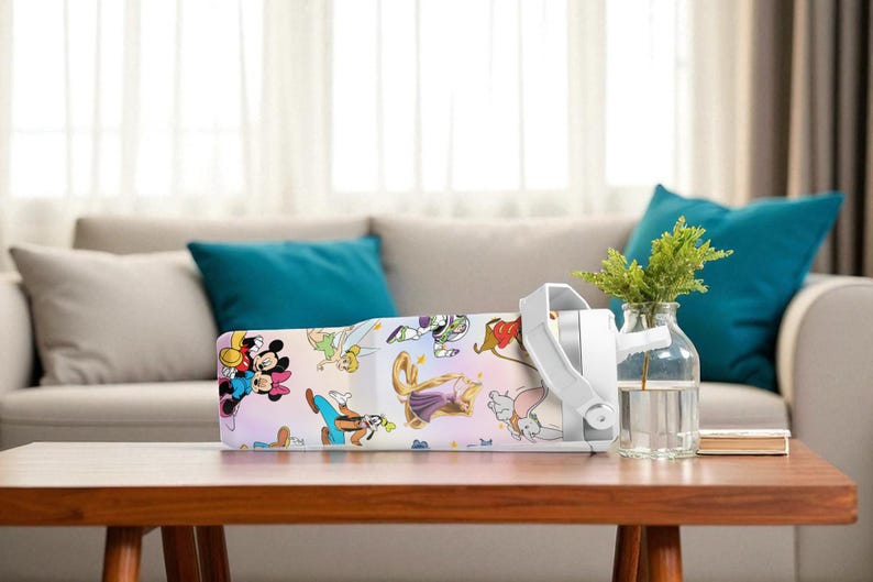 May include: A white air purifier featuring Disney characters, placed on a wooden table. It has a white handle and sits beside a glass vase with green plants. The background shows a sofa.