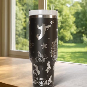 May include: Black stainless steel tumbler with a white lid and straw. The tumbler features silver illustrations of characters and symbols from "The Little Mermaid," including Ariel, King Triton, and a starfish. The text "THE LITTLE MERMAID" is also visible.