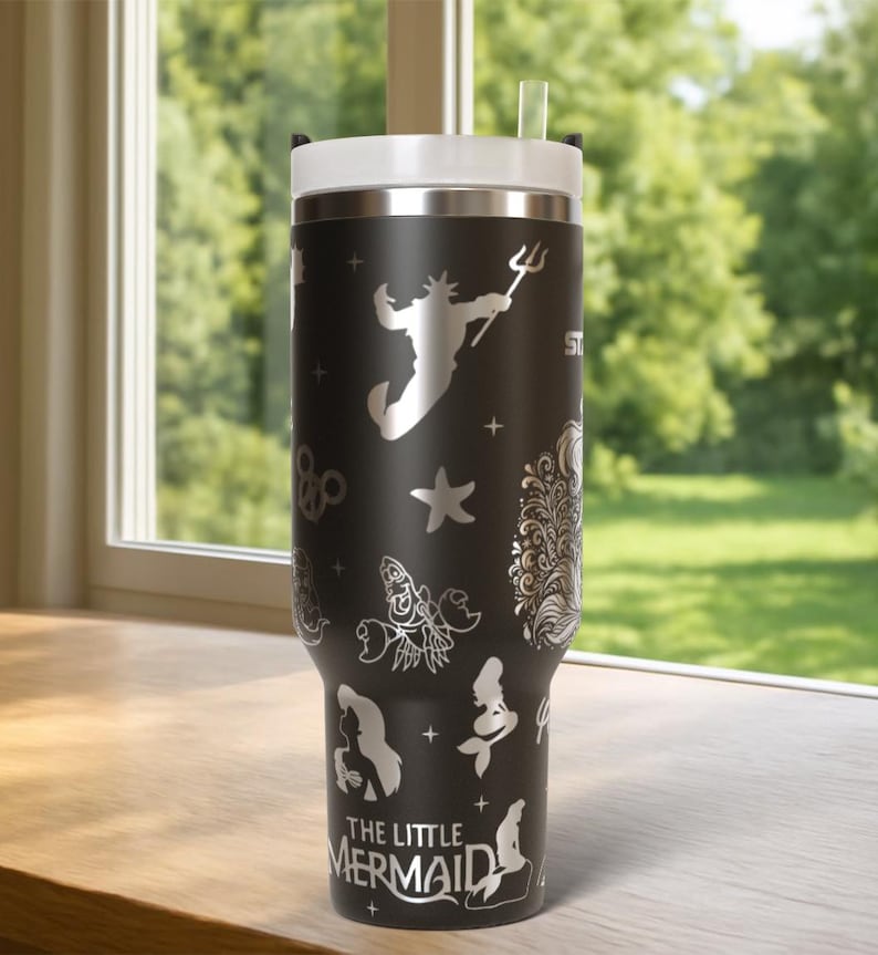 May include: Black stainless steel tumbler with a white lid and straw. The tumbler features silver illustrations of characters and symbols from "The Little Mermaid," including Ariel, King Triton, and a starfish. The text "THE LITTLE MERMAID" is also visible.