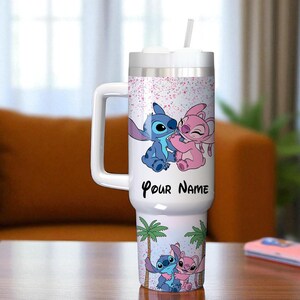 May include: A white tumbler with a handle, featuring a cartoon design of a blue and pink alien couple. The tumbler has a white lid and straw, with the text "Your Name" on the front. The design includes palm trees and a pink speckled background.