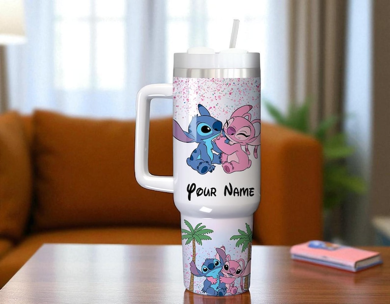 May include: A white tumbler with a handle, featuring a cartoon design of a blue and pink alien couple. The tumbler has a white lid and straw, with the text "Your Name" on the front. The design includes palm trees and a pink speckled background.