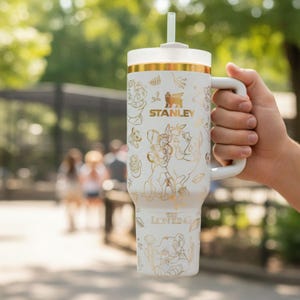 May include: White Stanley travel mug with gold detailing and a handle, featuring gold illustrations of "The Lion King" characters. The mug has a white lid and straw, with the "Stanley" logo visible.