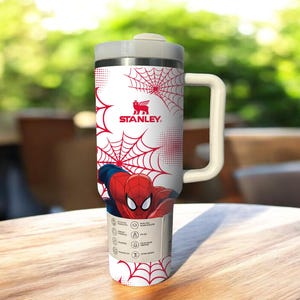 May include: A white Stanley travel mug featuring a red spiderweb design and a graphic of Spider-Man. The mug has a gray lid and handle. The Stanley logo is visible. The mug is on a wooden table.