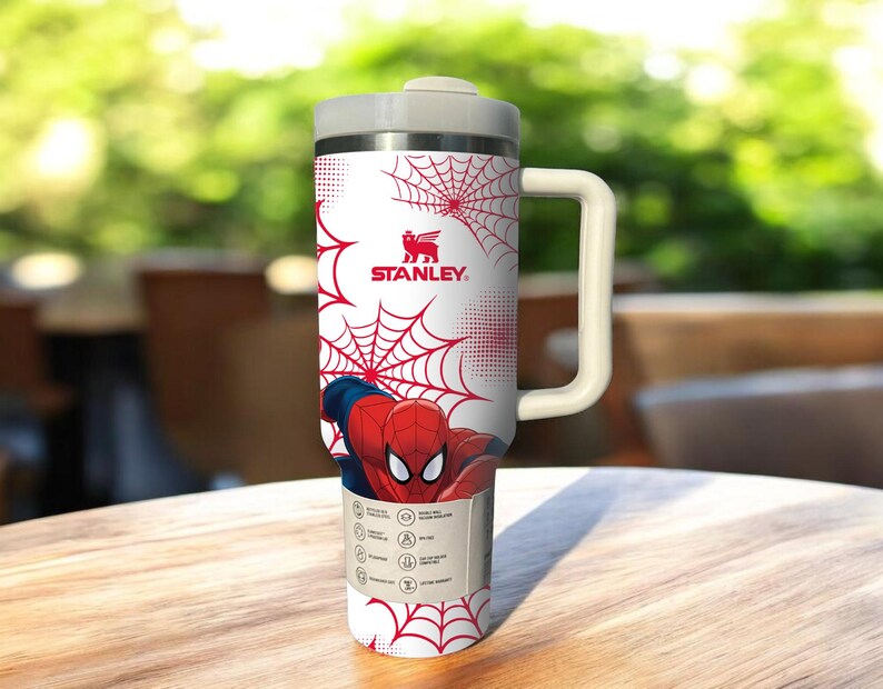 May include: A white Stanley travel mug featuring a red spiderweb design and a graphic of Spider-Man. The mug has a gray lid and handle. The Stanley logo is visible. The mug is on a wooden table.
