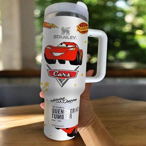 May include: White Stanley Quencher H2.0 Flowstate tumbler featuring Lightning McQueen from the Cars movie. The tumbler has a handle and a lid. The design includes the Cars logo, the Stanley logo, and the text "All Day Hydration."
