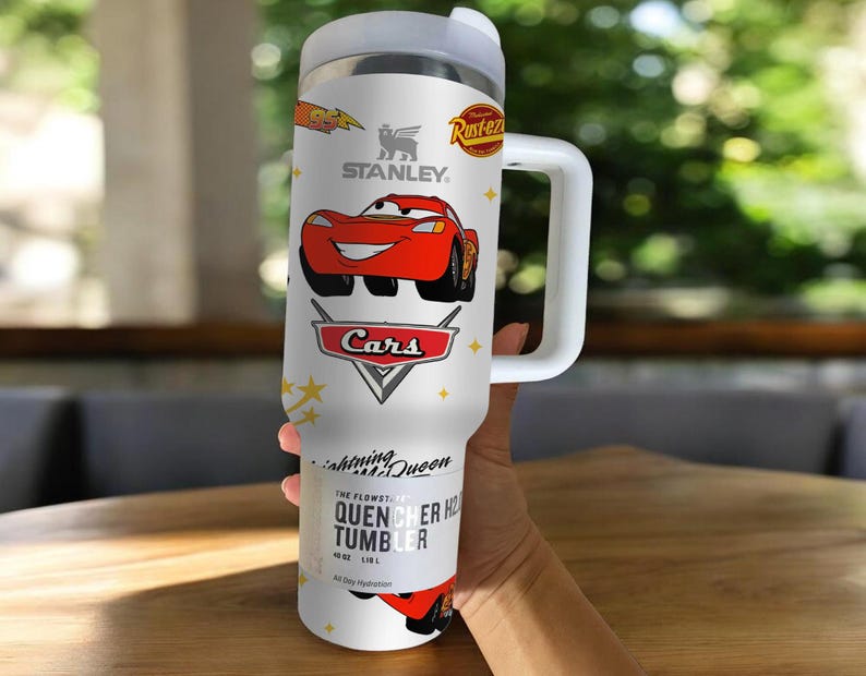 May include: White Stanley Quencher H2.0 Flowstate tumbler featuring Lightning McQueen from the Cars movie. The tumbler has a handle and a lid. The design includes the Cars logo, the Stanley logo, and the text "All Day Hydration."