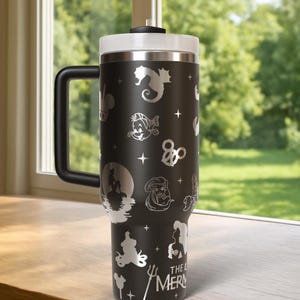 May include: Black travel mug with a white lid and handle. The mug features etched images of characters and symbols from "The Little Mermaid," including Ariel, Flounder, and a seahorse. The text "THE LITTLE MERMAID" is also etched on the mug.