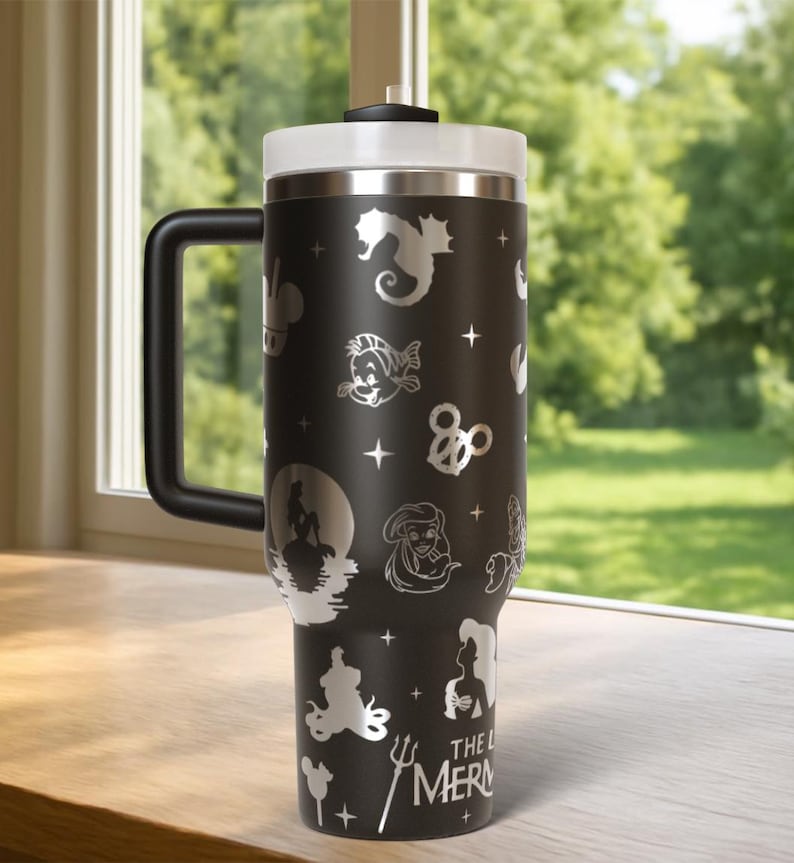 May include: Black travel mug with a white lid and handle. The mug features etched images of characters and symbols from "The Little Mermaid," including Ariel, Flounder, and a seahorse. The text "THE LITTLE MERMAID" is also etched on the mug.