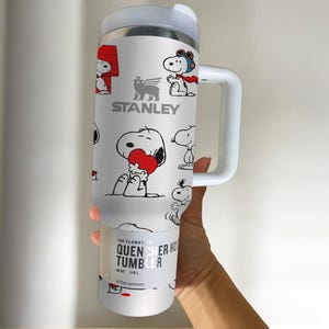 May include: White Stanley Quencher H2.0 Flowstate tumbler featuring Snoopy cartoon graphics. The tumbler has a white handle and lid. The text "STANLEY" is visible. The bottom of the tumbler reads "QUENCHER H2.0 TUMBLER" and "40 oz / 1.18 L".
