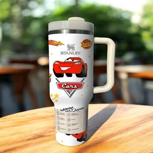 May include: White Stanley travel mug featuring a red cartoon car design from the movie Cars. The mug has a handle, a gray lid, and the Stanley logo. The text "Lightning McQueen" is also visible.