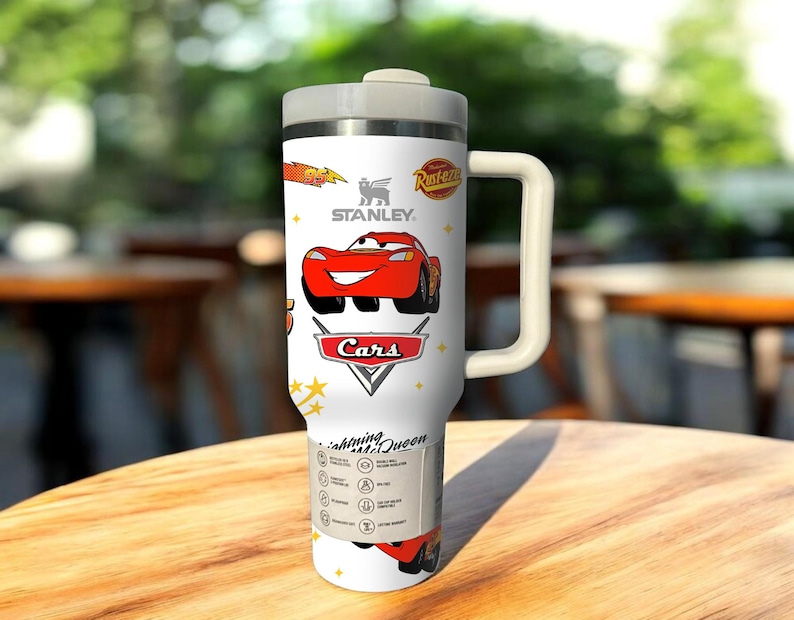 May include: White Stanley travel mug featuring a red cartoon car design from the movie Cars. The mug has a handle, a gray lid, and the Stanley logo. The text "Lightning McQueen" is also visible.