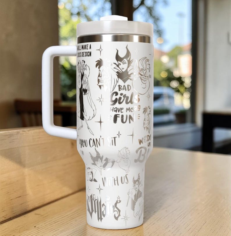 Puede incluir: Vaso t&eacute;rmico blanco con asa, con ilustraciones de villanos de Disney y el texto "BAD Girls HAVE MORE FUN". El vaso tiene tapa y asa blancas, para bebidas fr&iacute;as o calientes.