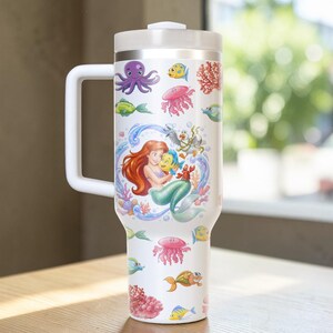 Puede incluir: Vaso t&eacute;rmico blanco con asa, con un dise&ntilde;o colorido de Ariel la sirenita, Flounder y varias criaturas marinas. El dise&ntilde;o incluye un pulpo, medusas y coral. El vaso tiene tapa y asa blancas.
