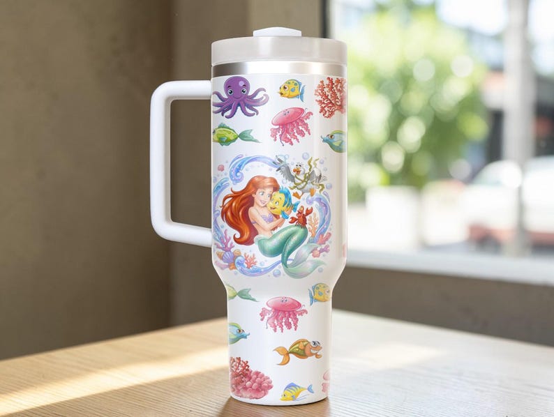 Puede incluir: Vaso t&eacute;rmico blanco con asa, con un dise&ntilde;o colorido de Ariel la sirenita, Flounder y varias criaturas marinas. El dise&ntilde;o incluye un pulpo, medusas y coral. El vaso tiene tapa y asa blancas.