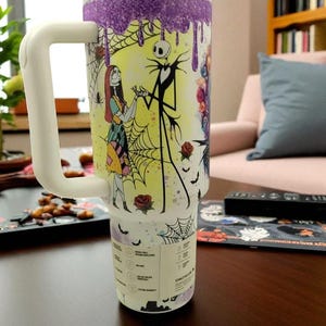 May include: A large white travel mug with a handle, featuring a colorful design of Jack Skellington and Sally from The Nightmare Before Christmas. The mug has a white lid and a purple glitter drip design at the top.
