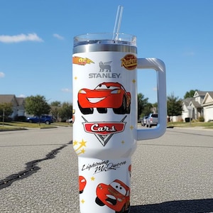May include: A white Stanley tumbler featuring Lightning McQueen from the Cars movie. The tumbler has a handle, a clear lid, and a straw. The design includes the Cars logo, the number 95, and the words "Lightning McQueen".