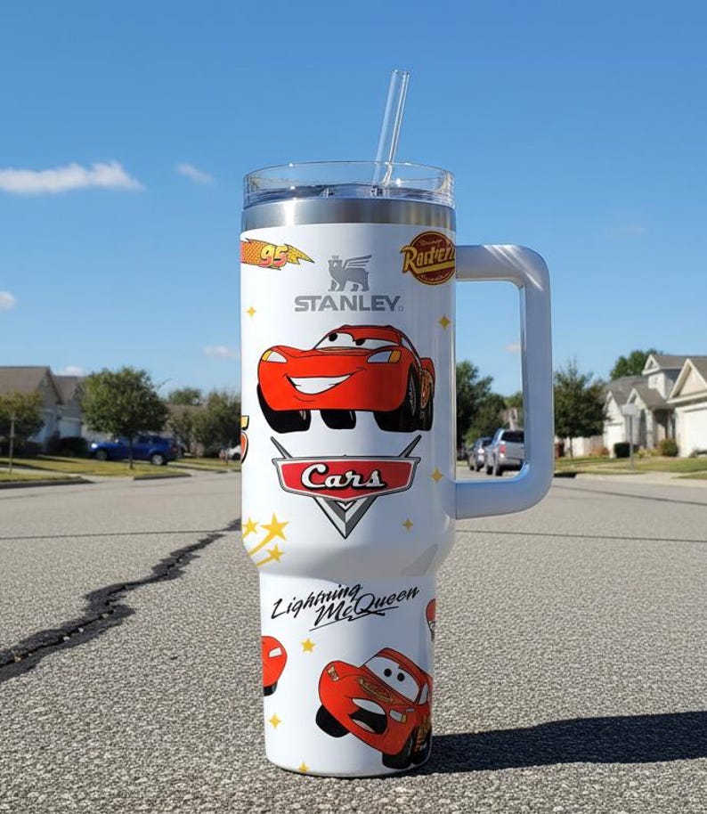 May include: A white Stanley tumbler featuring Lightning McQueen from the Cars movie. The tumbler has a handle, a clear lid, and a straw. The design includes the Cars logo, the number 95, and the words "Lightning McQueen".