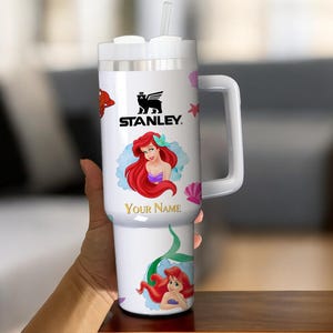 Puede incluir: Vaso Stanley blanco con asa, pajita y tapa. El vaso est&aacute; decorado con una imagen de dibujos animados de Ariel de La Sirenita, el texto "Your Name" y el logotipo de Stanley. Tambi&eacute;n se presentan conchas marinas rosas y otras criaturas marinas.