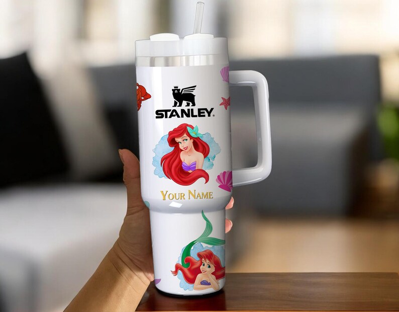 Puede incluir: Vaso Stanley blanco con asa, pajita y tapa. El vaso est&aacute; decorado con una imagen de dibujos animados de Ariel de La Sirenita, el texto "Your Name" y el logotipo de Stanley. Tambi&eacute;n se presentan conchas marinas rosas y otras criaturas marinas.