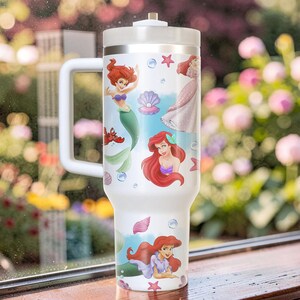 Puede incluir: Vaso t&eacute;rmico blanco con ilustraciones de Ariel de La Sirenita. El vaso tiene un asa y una tapa blancas, con im&aacute;genes de Ariel, conchas marinas y estrellas de mar. El vaso est&aacute; dise&ntilde;ado para contener una gran cantidad de l&iacute;quido.