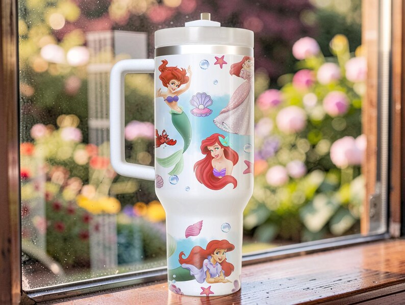 Puede incluir: Vaso t&eacute;rmico blanco con ilustraciones de Ariel de La Sirenita. El vaso tiene un asa y una tapa blancas, con im&aacute;genes de Ariel, conchas marinas y estrellas de mar. El vaso est&aacute; dise&ntilde;ado para contener una gran cantidad de l&iacute;quido.