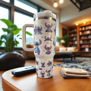 May include: A white travel mug featuring a repeating pattern of the character Stitch from the Disney movie Lilo & Stitch. The mug has a white handle and lid, and is sitting on a wooden table. A remote control and coasters are also on the table.