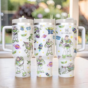 Pu&ograve; includere: Tre bicchieri Stanley bianchi con manici, adornati con illustrazioni colorate di Buzz Lightyear e altri personaggi di Toy Story. I bicchieri hanno un coperchio bianco e una cannuccia. Il logo Stanley &egrave; visibile su uno dei bicchieri.