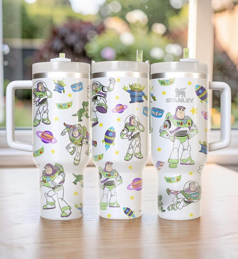 Pu&ograve; includere: Tre bicchieri Stanley bianchi con manici, adornati con illustrazioni colorate di Buzz Lightyear e altri personaggi di Toy Story. I bicchieri hanno un coperchio bianco e una cannuccia. Il logo Stanley &egrave; visibile su uno dei bicchieri.