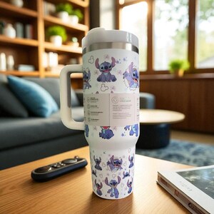 May include: A white insulated tumbler with a handle, featuring a repeating pattern of the character Stitch from Lilo & Stitch. The tumbler has a stainless steel top and a white handle. The design includes Stitch illustrations in various poses, with pink flowers.