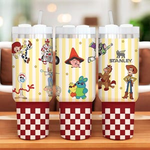 May include: Three stainless steel insulated tumblers with white handles and lids. The tumblers feature a yellow and white striped pattern with various Toy Story character graphics. The base is red and white checkered. The Stanley logo is visible.