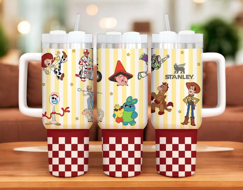 May include: Three stainless steel insulated tumblers with white handles and lids. The tumblers feature a yellow and white striped pattern with various Toy Story character graphics. The base is red and white checkered. The Stanley logo is visible.