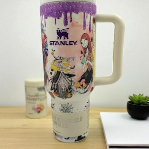 May include: A white Stanley Quencher H2.0 Tumbler with a purple dripped design at the top. The tumbler features colorful illustrations of characters from The Nightmare Before Christmas. It has a white handle and a lid.