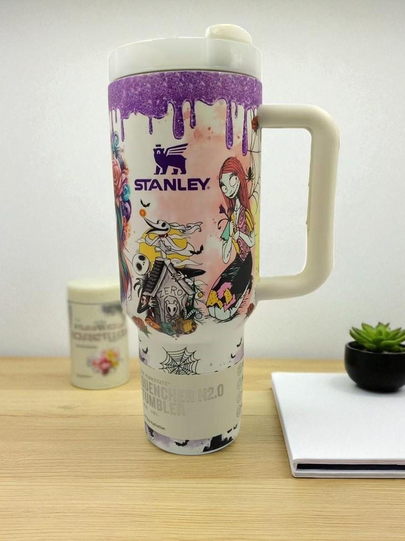 May include: A white Stanley Quencher H2.0 Tumbler with a purple dripped design at the top. The tumbler features colorful illustrations of characters from The Nightmare Before Christmas. It has a white handle and a lid.