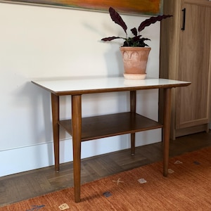May include: A rectangular side table with a smooth white top and a lower wooden shelf. The table features four tapered wooden legs and a warm brown finish on the frame. A terracotta pot containing a dark-leafed plant sits on the table surface.