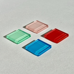 Puede incluir: Cuatro piezas cuadradas de material translúcido, cada una con un color distinto: rosa claro, verde brillante, rojo vibrante y azul intenso. Cada pieza tiene una superficie lisa y plana con un borde ligeramente elevado en uno de sus lados, creando una forma geométrica uniforme.