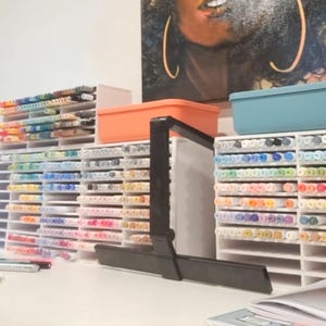 May include: A selection of colourful art markers arranged in white tiered storage units. The markers are sorted by colour, with a variety of shades visible. An orange and blue container are on top of the storage units. A black adjustable stand is in the foreground.