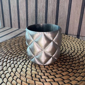 Peut inclure: Un récipient cylindrique argenté matelassé. Le récipient présente un motif en losanges et une base arrondie. L'objet est posé sur un tapis circulaire tissé. L'arrière-plan est un mur en bois avec des rayures verticales.