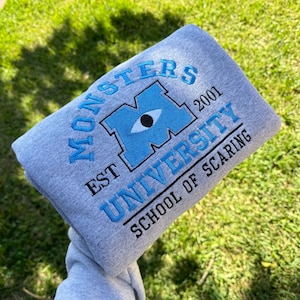 以下が含まれることがあります： グレーのトレーナーに、青い刺繍で「モンスターズ・ユニバーシティ」とロゴが入っています。「M」は青い目で、黒い瞳をしています。「Est 2001」という文字がロゴの上にあり、「School of Scaring」という文字がロゴの下にあります。