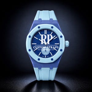 Könnte beinhalten: Eine hellblaue Armbanduhr mit dunkelblauem, texturiertem Zifferblatt und dem weißen Schriftzug RP, CUSTOM STRAPS, LONDON und Royal Pop. Die Uhr hat eine hellblaue achteckige Lünette mit acht sichtbaren Schrauben und ein passendes Kautschukarmband.